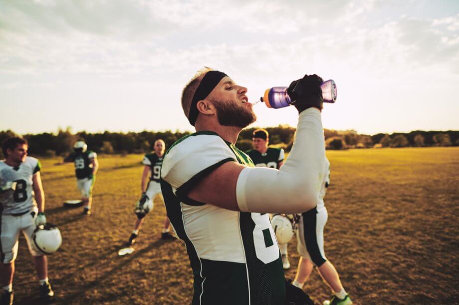 Photograph of a male football player on a football field at dusk is drinking a purple unnamed sports drink.