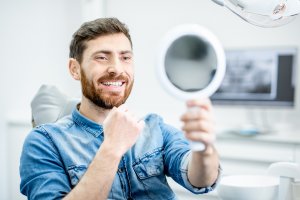 Photograph of a man wearing a blue shirt, smiling while looking at himself in a handheld mirror while at a dentist office.