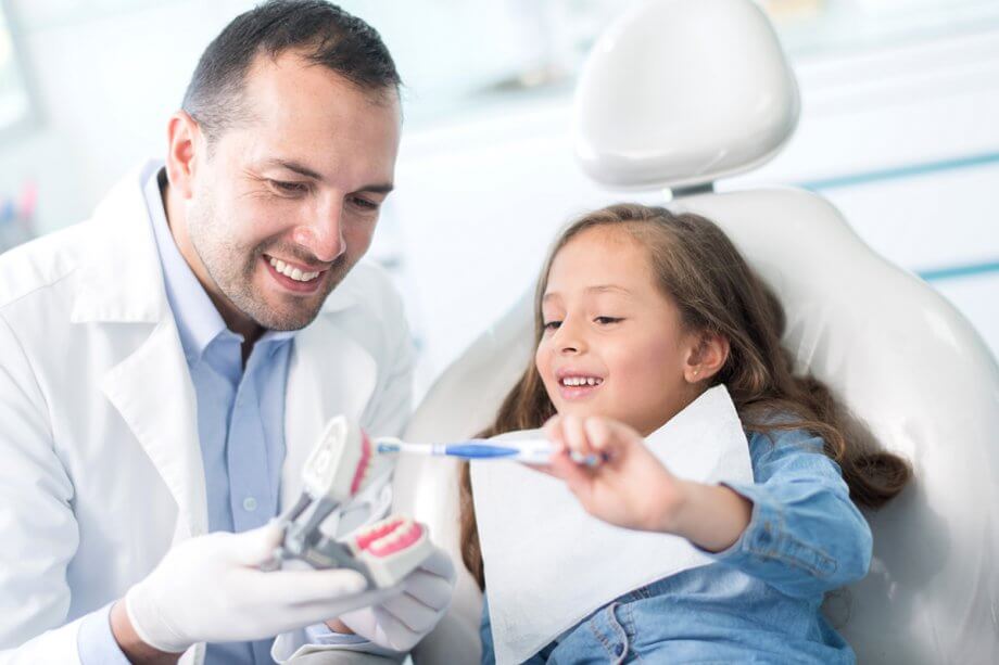 young girl in exam chair with dentist holding model of teeth
