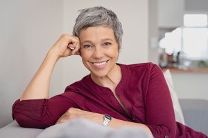 older woman in burgundy shirt, sitting on couch, smiling